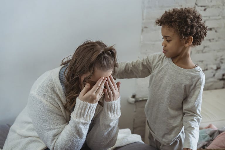 Caring African American son touching shoulder of upset faceless mother covering face while sitting in light room near wall at home